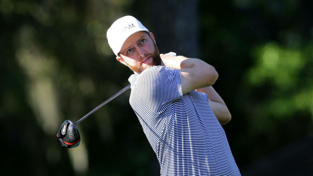 Golfer Chris Kirk mid-swing focused ahead in striped shirt and white cap on green golf course Wallpaper