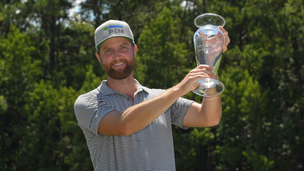 Chris Kirk holding clear glass trophy outdoors wearing striped polo and gray cap with green trees background Wallpaper