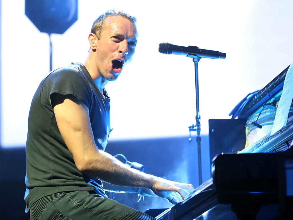 Chris Martin performing live at Glastonbury Festival on piano with dramatic blue white lighting Wallpaper