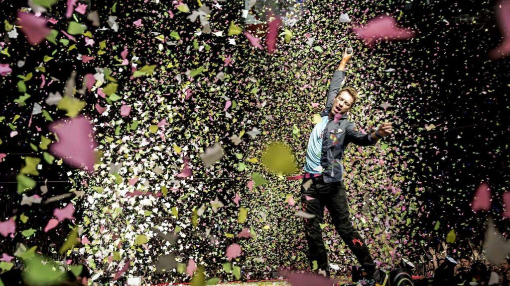 Chris Martin of Coldplay center stage with vibrant pink green yellow confetti at live concert Wallpaper