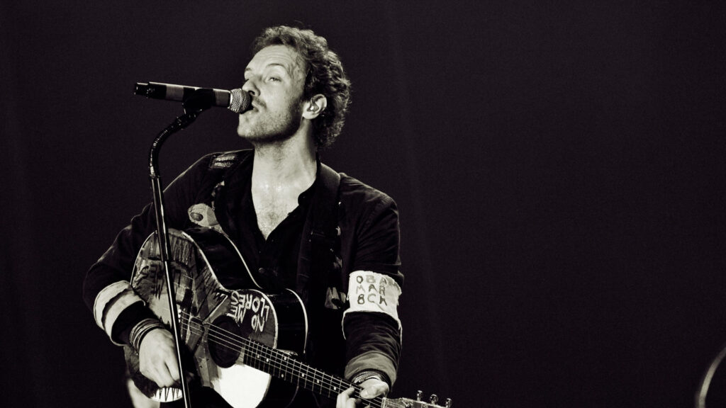 Chris Martin of Coldplay passionately sings and plays guitar in black and white concert wallpaper
