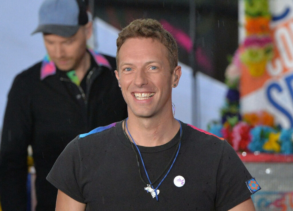 Chris Martin from Coldplay smiling warmly at Rockefeller Plaza with vibrant colorful decorations Wallpaper