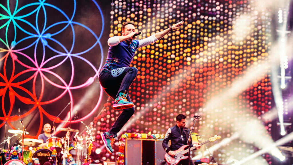 Chris Martin jumping energetically at Coldplay concert with colorful lights and band members Wallpaper
