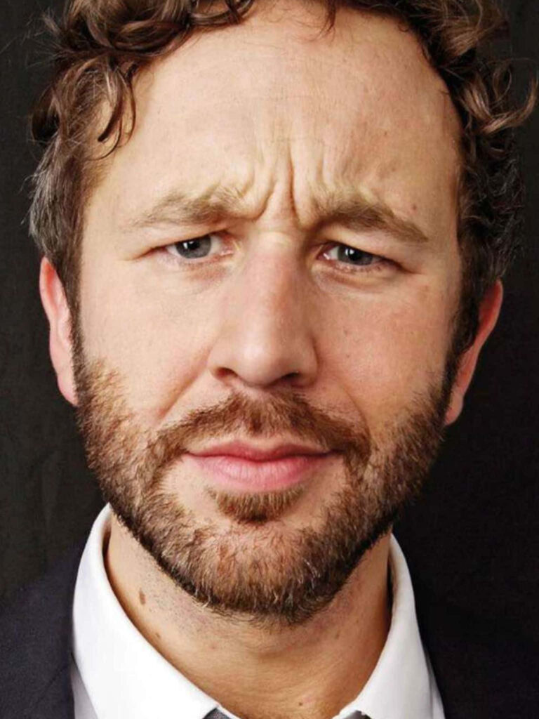 Close-up portrait of Chris O'Dowd with curly hair and beard against dark wallpaper background