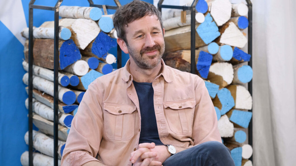 Chris O'Dowd seated by vibrant birch logs with blue white accents showcasing casual style background Wallpaper