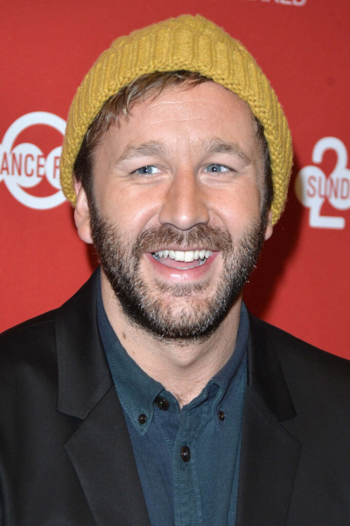 Chris O'Dowd smiling in mustard yellow beanie and dark jacket on vibrant red wallpaper background