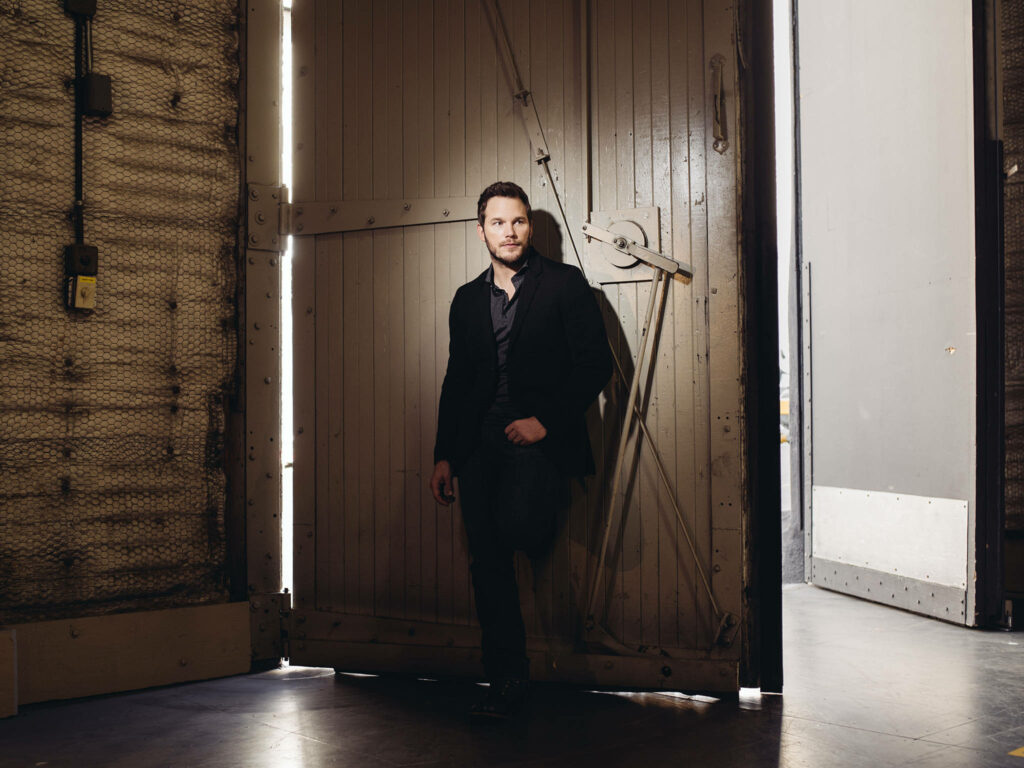 Chris Pratt in dark suit posing confidently by rustic wooden door with warm dramatic lighting Wallpaper