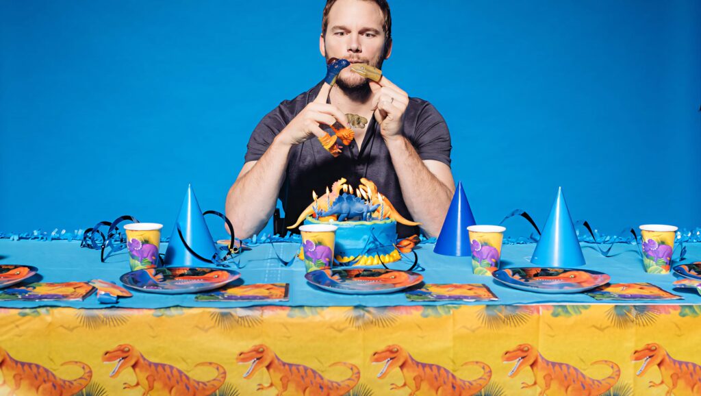 Chris Pratt at vibrant Jurassic-themed party table with colorful dinosaur plates cups and hats Wallpaper