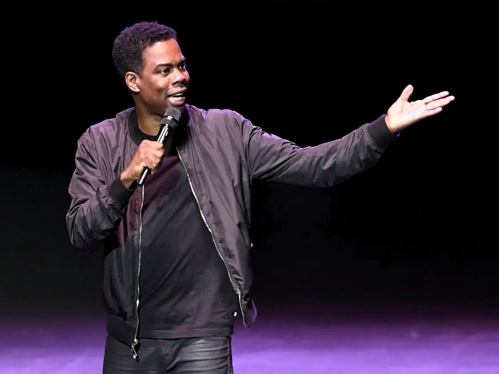 Comedian Chris Rock performing stand-up on stage with microphone against dark background wallpaper