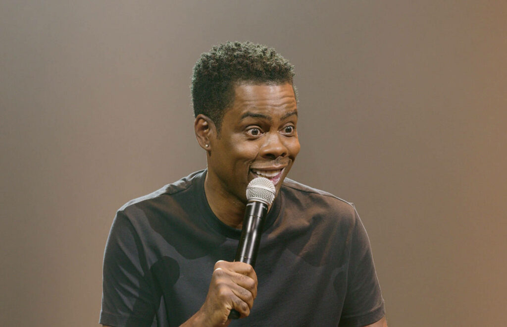 Comedian Chris Rock on stage with microphone playful expression soft lighting wallpaper background