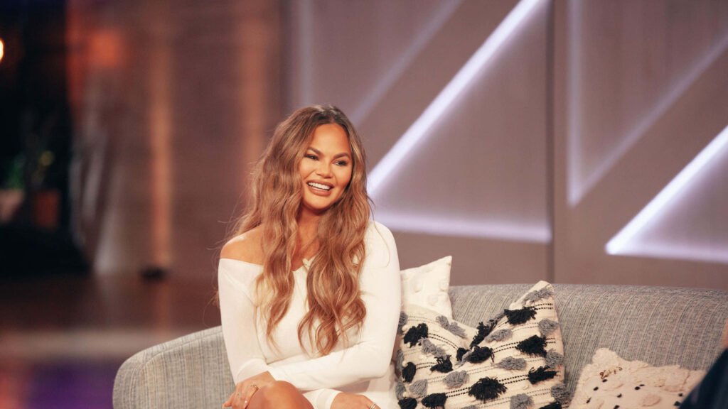 Chrissy Teigen Smiles in Elegant Off-Shoulder Outfit on Cozy Sofa Wallpaper