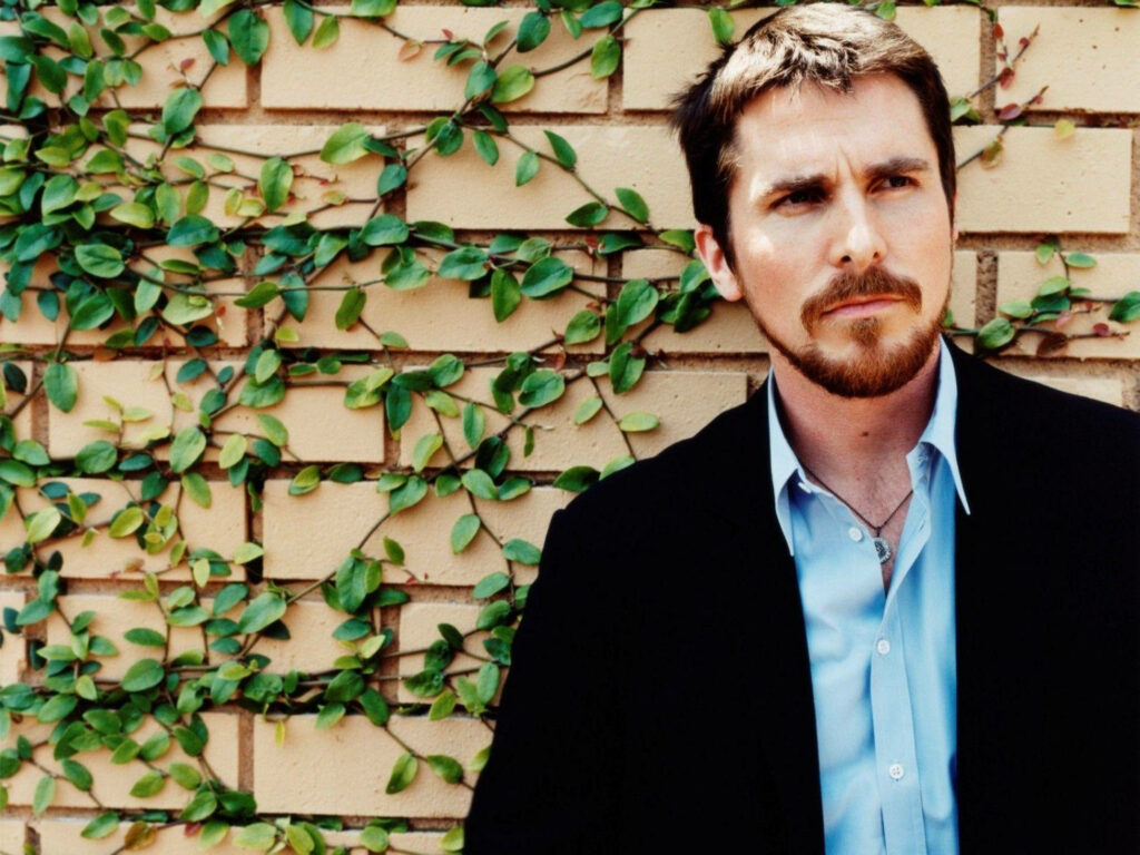 Christian Bale in blue shirt and black jacket stands confidently against vine-covered brick wall wallpaper