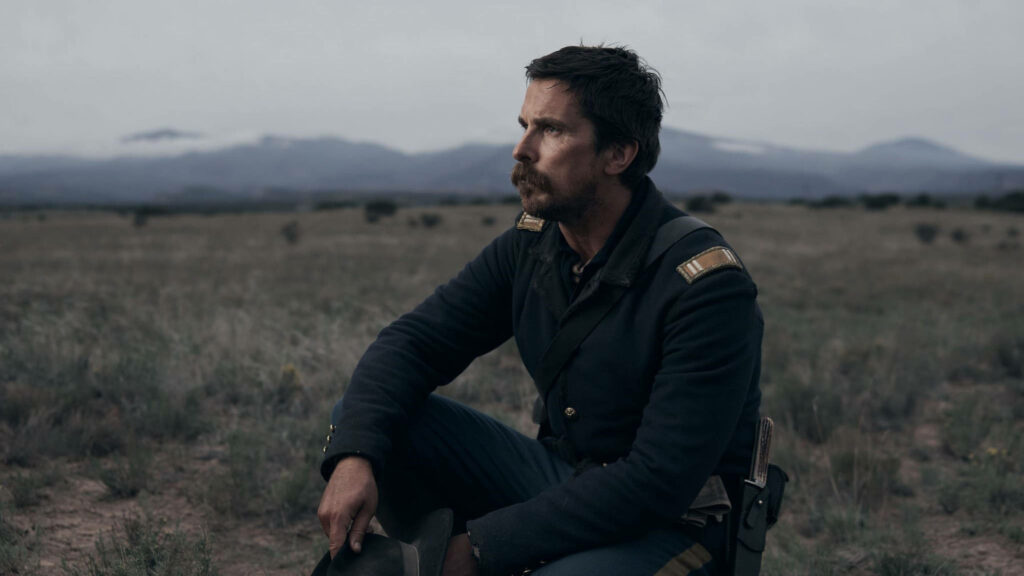 Christian Bale as military officer in Hostiles 2017 sitting thoughtfully in rugged mountain landscape Wallpaper