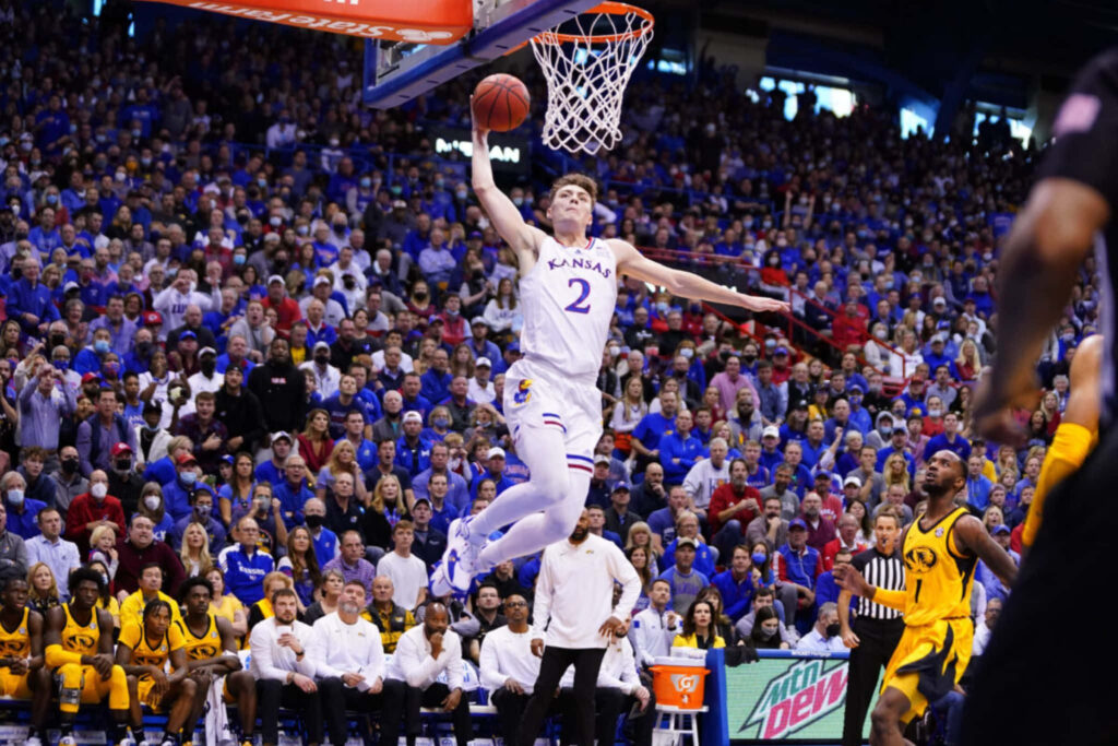 Christian Braun mid dunk in Kansas basketball uniform during intense game with vibrant crowd Wallpaper