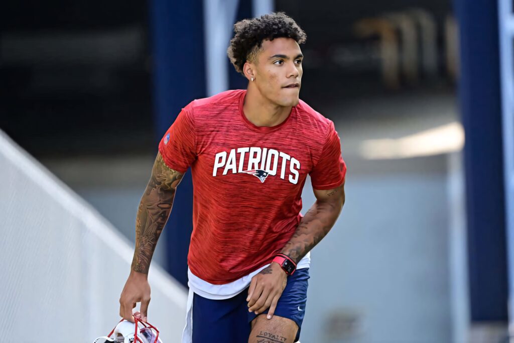 Christian Gonzalez in red Patriots shirt holding helmet showing tattoos and focused expression Wallpaper