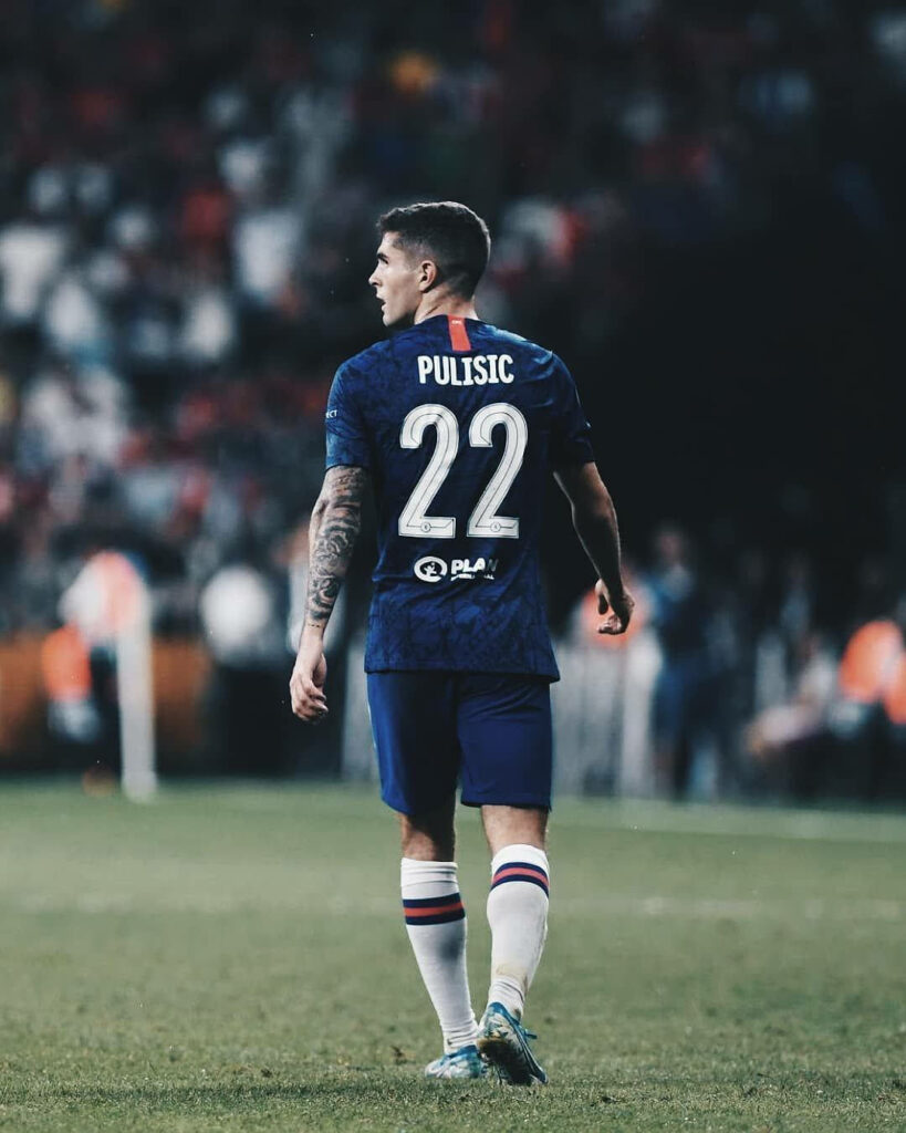 Christian Pulisic in number 22 jersey focused on field with blurred stadium and blue tones background Wallpaper
