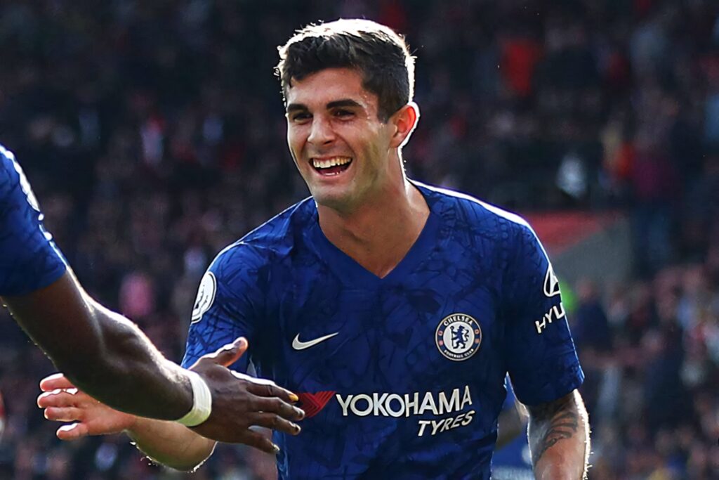 Christian Pulisic smiling in blue Chelsea jersey on football field with blurred stadium background Wallpaper