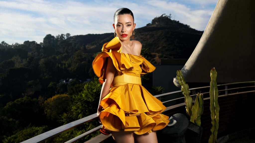 Christian Serratos in yellow ruffled dress on balcony overlooking scenic hillside natural landscape Wallpaper
