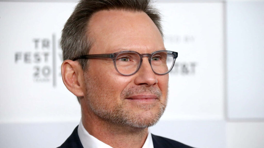 Christian Slater in formal suit with signature glasses smiling against soft neutral background wallpaper