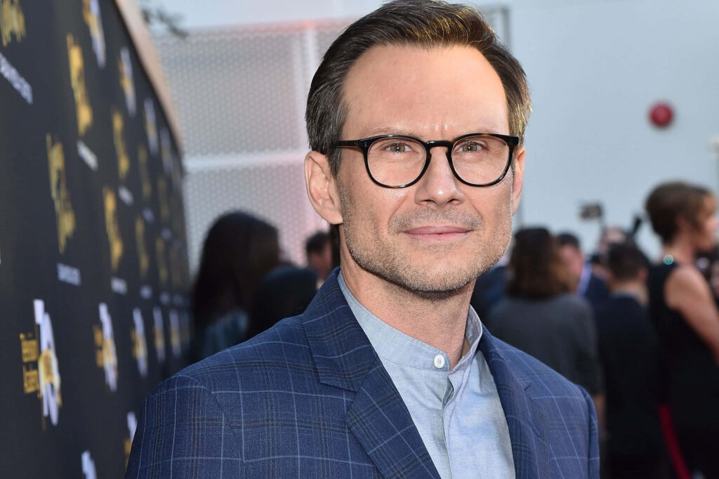 Christian Slater in blue checkered suit and glasses at stylish event with blurred crowd background Wallpaper