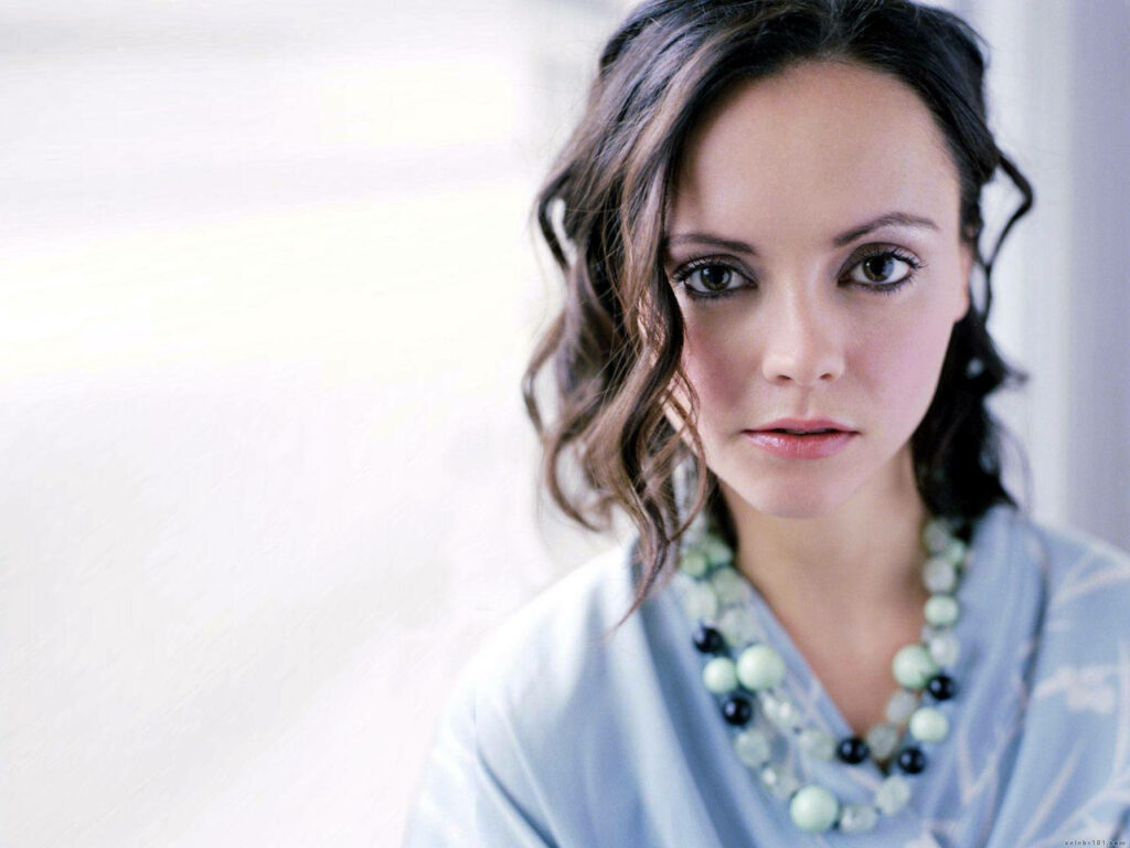 Christina Ricci in pastel outfit with statement bead necklace softly lit against wallpaper background