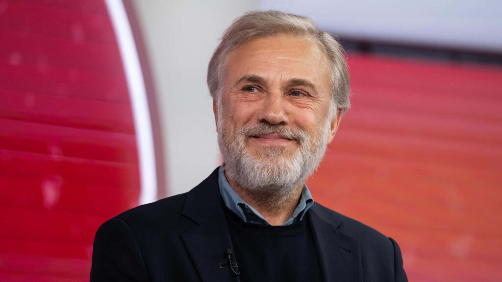 Christoph Waltz portrait with warm smile salt and pepper beard elegant attire red orange background Wallpaper