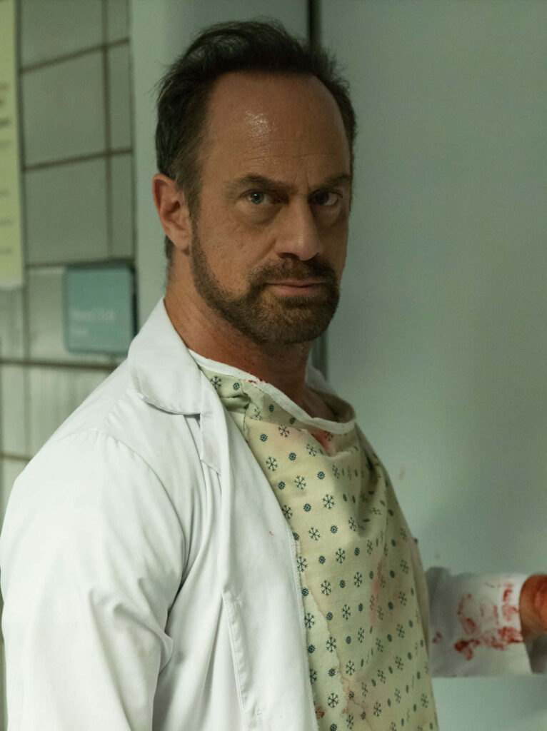 Christopher Meloni in blood-stained hospital gown and white coat in dimly lit tiled room wallpaper