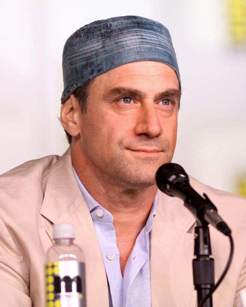 Christopher Meloni in light jacket and blue cap speaking at panel event with soft lighting background Wallpaper