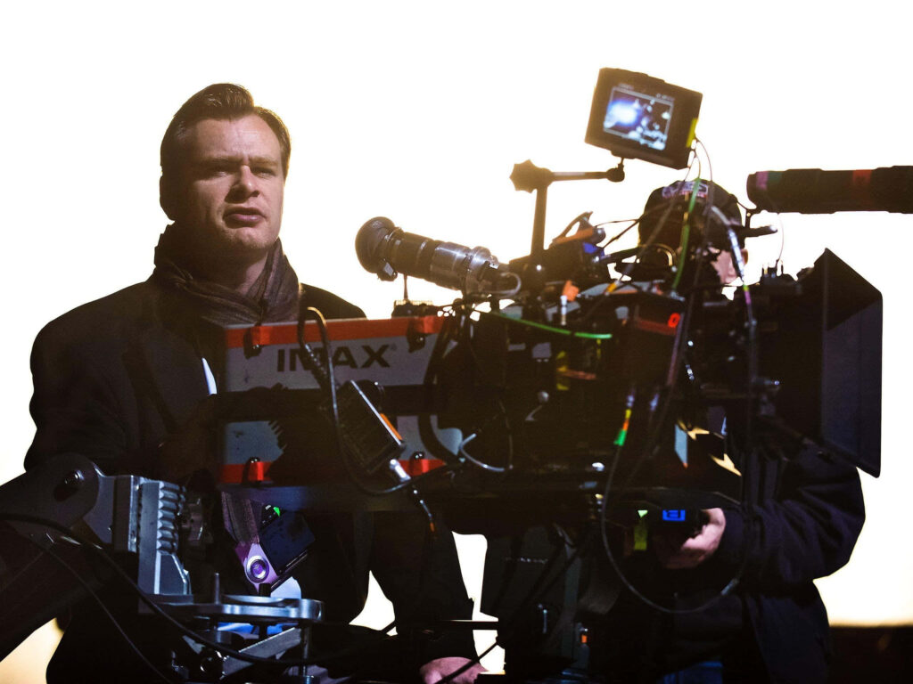 Christopher Nolan beside IMAX camera on set with dramatic lighting showcasing filmmaking focus Wallpaper