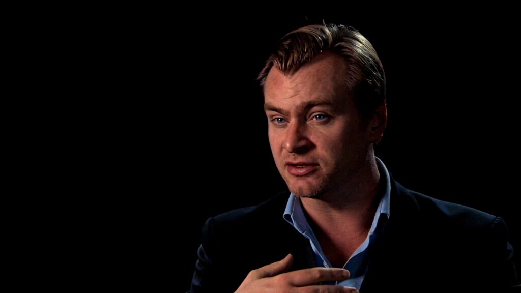 Christopher Nolan in formal attire engaging thoughtfully on film set with dark cinematic background Wallpaper