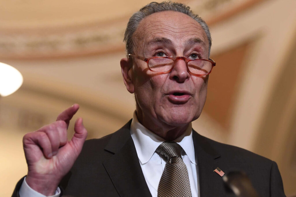 Chuck Schumer in a formal suit with thoughtful expression and warm lighting wallpaper background