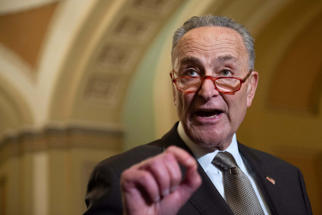 Chuck Schumer speaking in suit with red glasses framed by classic arched architecture wallpaper