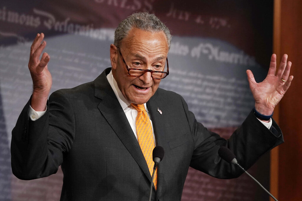 Chuck Schumer speaking at podium with raised hands and Constitution text background in suit and orange tie Wallpaper