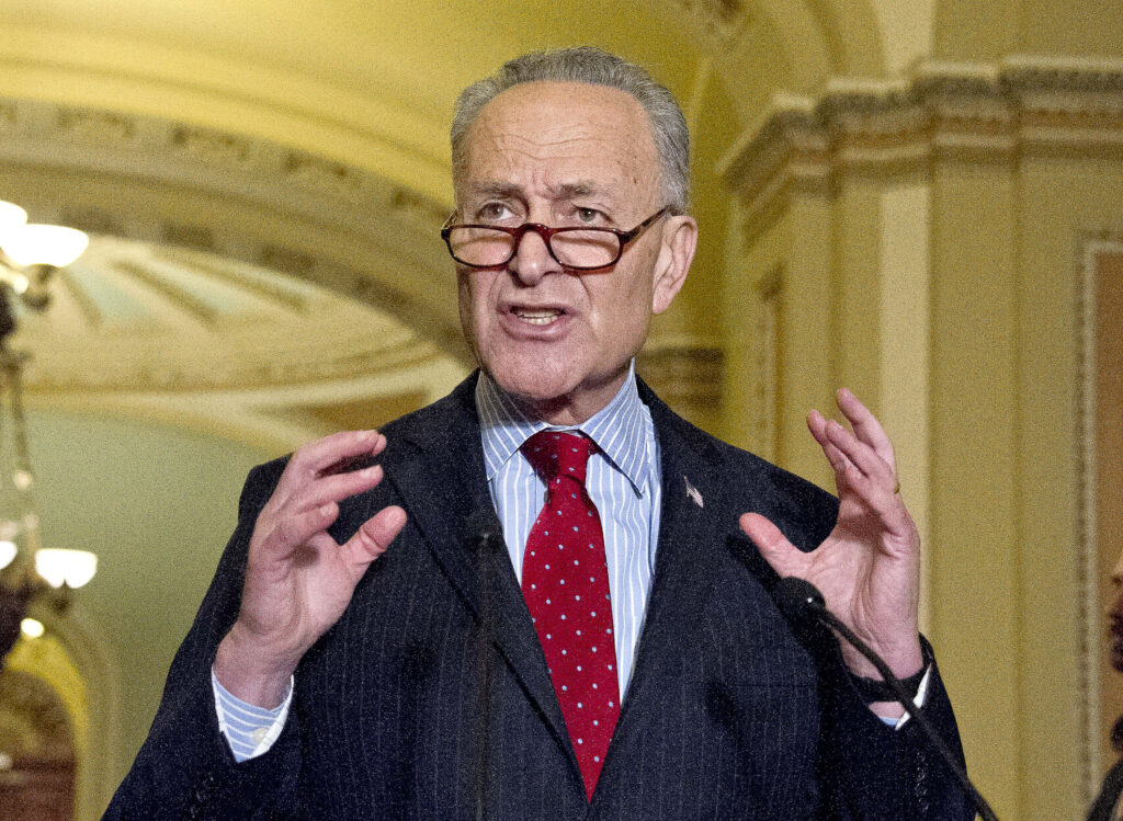 Chuck Schumer speaking in formal setting with warm lighting and classic architecture background Wallpaper