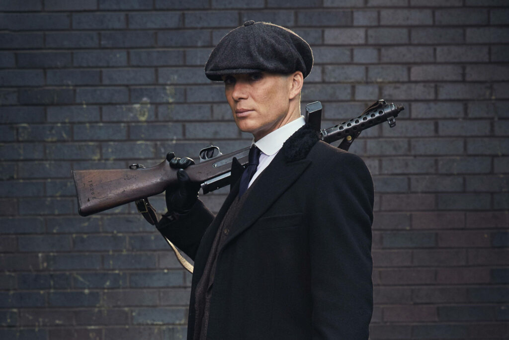 Cillian Murphy in flat cap and overcoat holding rifle against dark brick wall wallpaper background