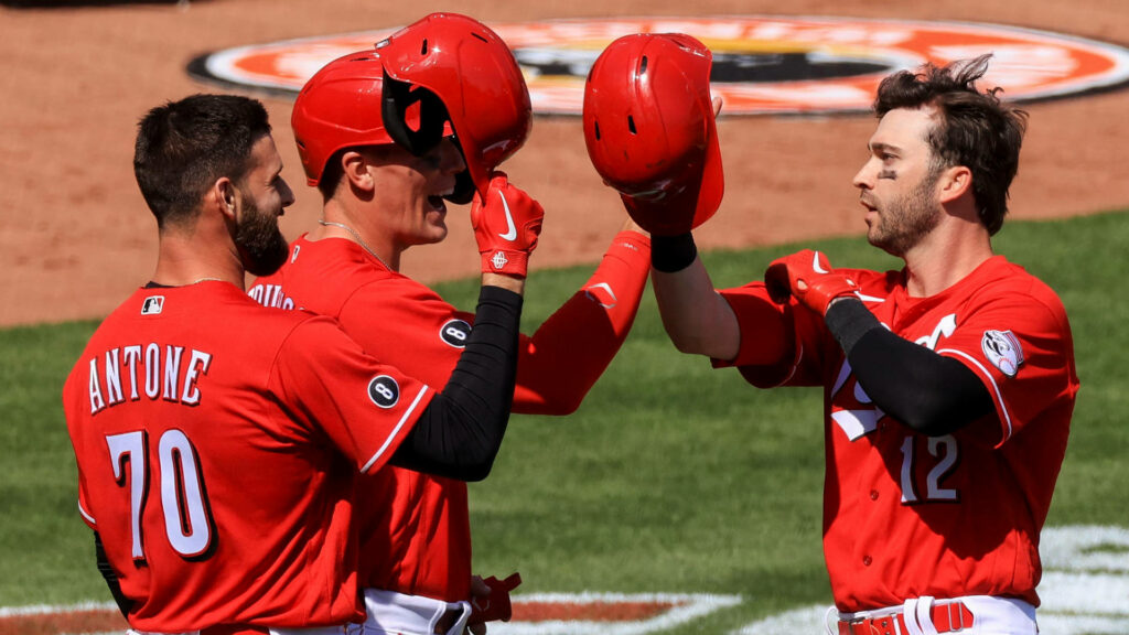 Three Cincinnati Reds players in red uniforms celebrate on field raising helmets in daylight victory Wallpaper