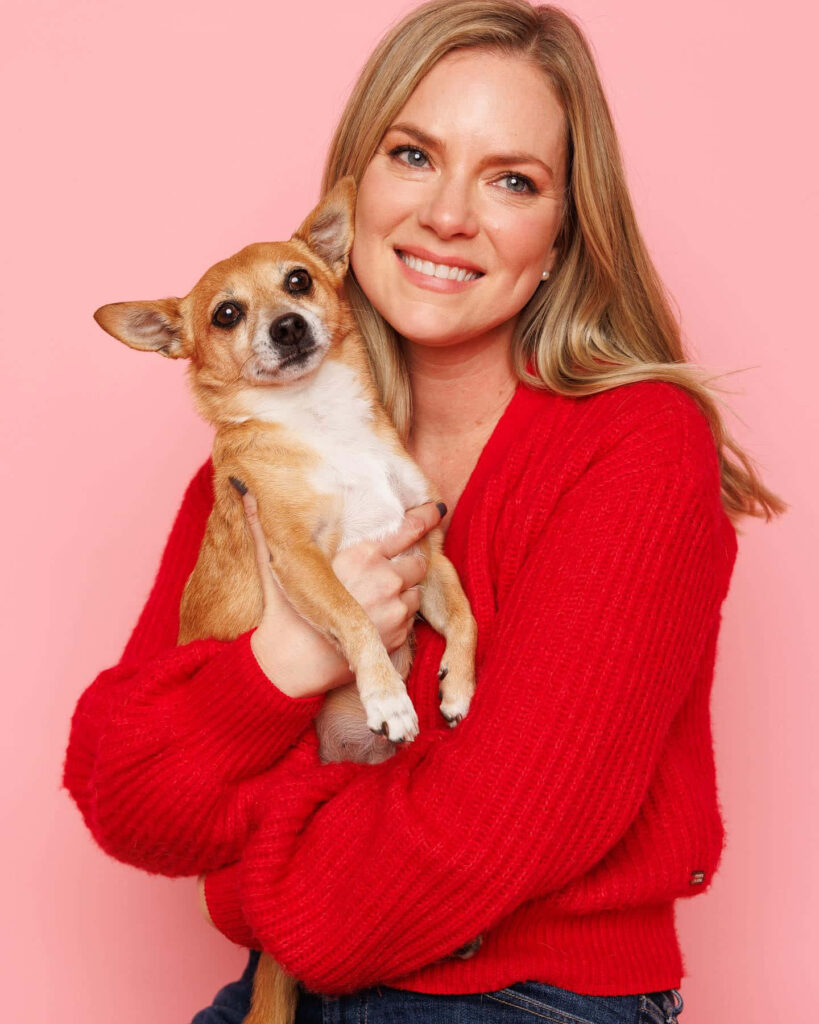 Cindy Busby smiling in red sweater holding small brown dog pink background cheerful wallpaper