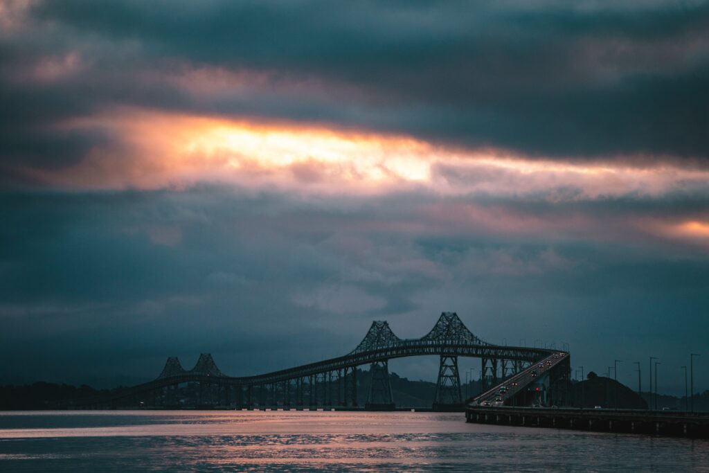 Long bridge over calm water at sunset with glowing car lights and dramatic colorful clouds Wallpaper