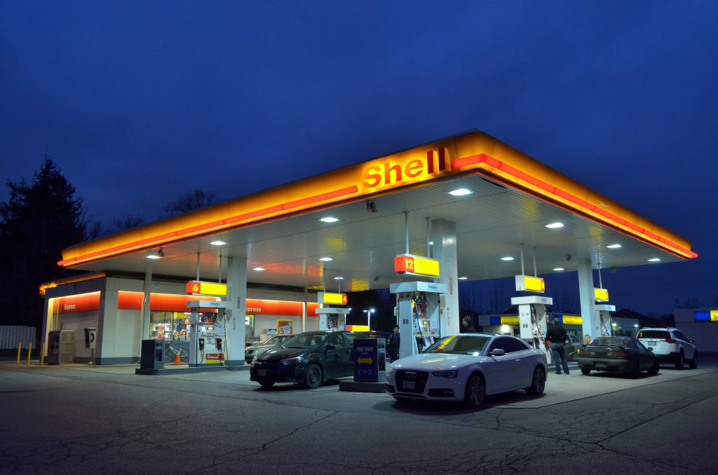 Shell gas station glowing yellow at twilight with fuel pumps and parked cars under dark sky Wallpaper