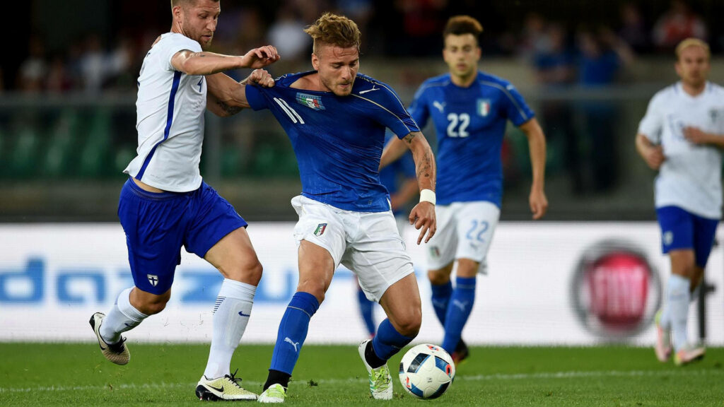 Ciro Immobile in blue Italy kit fiercely dribbling past defender during intense soccer match Wallpaper