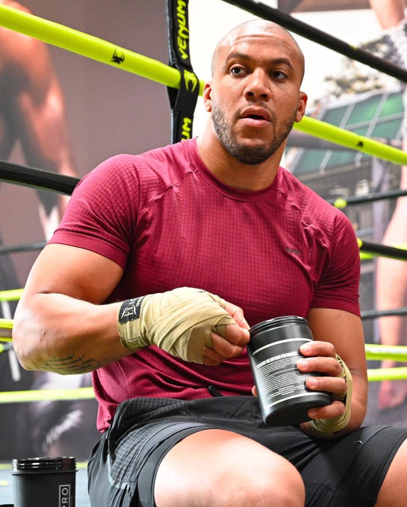 UFC heavyweight Ciryl Gane resting in boxing ring holding supplement tub in maroon shirt and wraps Wallpaper
