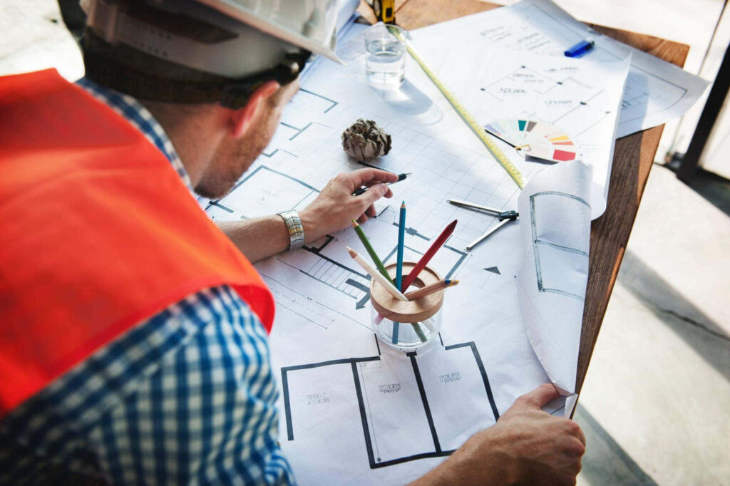 Civil engineer in safety vest reviewing detailed architectural blueprints with pencils and rulers Wallpaper