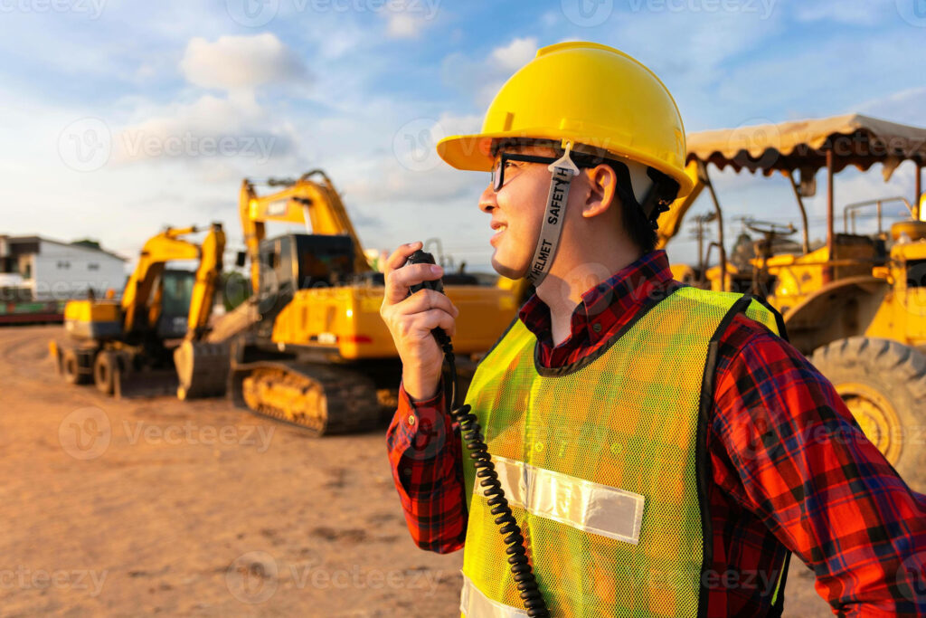 Civil engineer in yellow helmet and vest using radio at sunny construction site with machinery Wallpaper