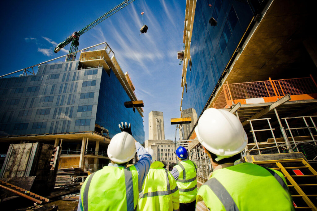 Construction workers in safety gear inspecting modern urban building site with crane and glass facades Wallpaper