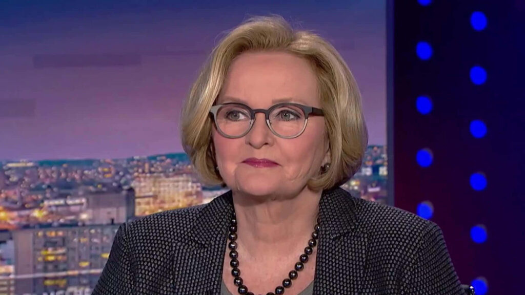 Claire McCaskill interview wearing glasses dark blazer beaded necklace blurred cityscape background Wallpaper