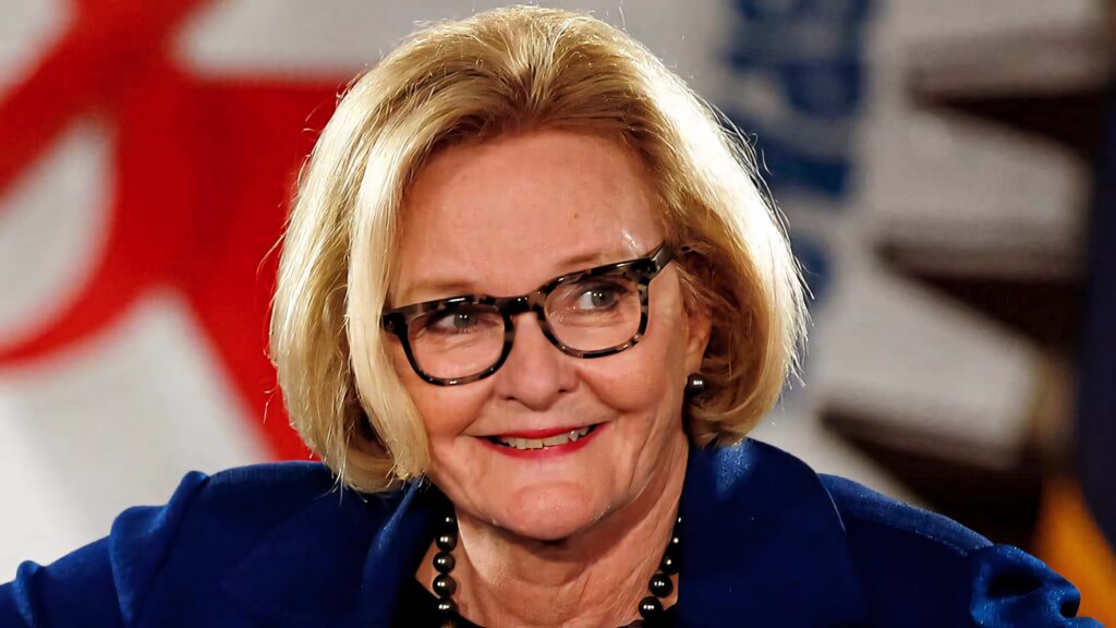 Claire McCaskill smiling in blue blazer and glasses with bright patterned professional background wallpaper