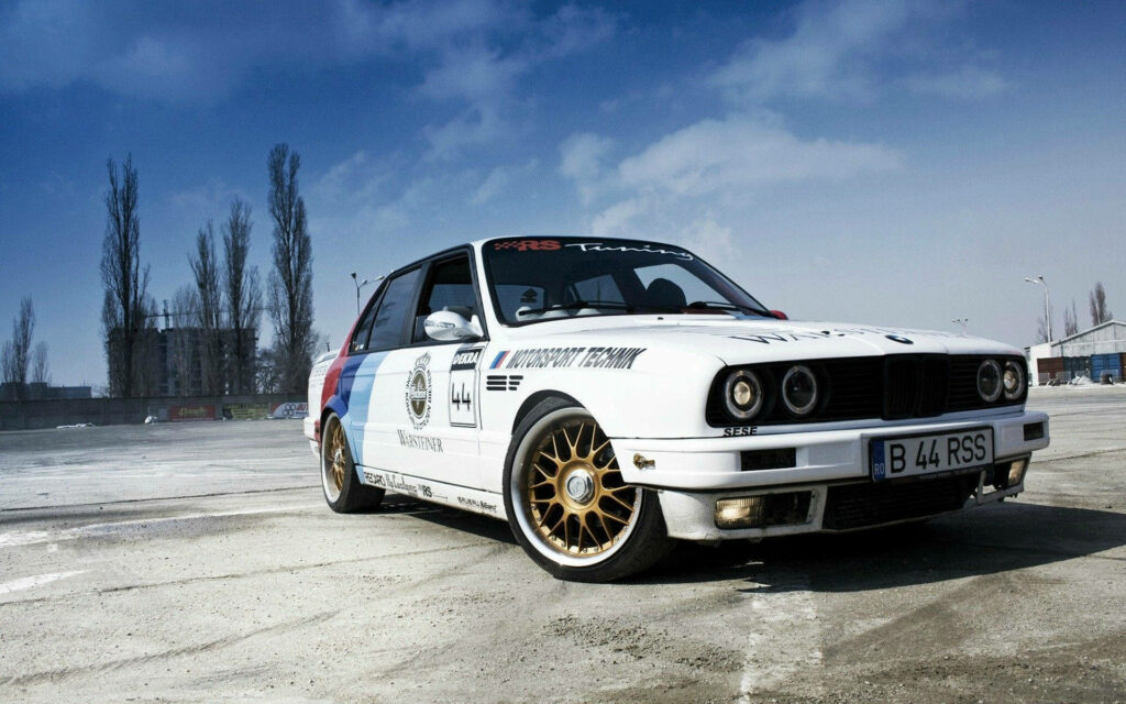 Classic BMW 1982-1994 with racing decals and gold wheels against blue sky urban backdrop wallpaper
