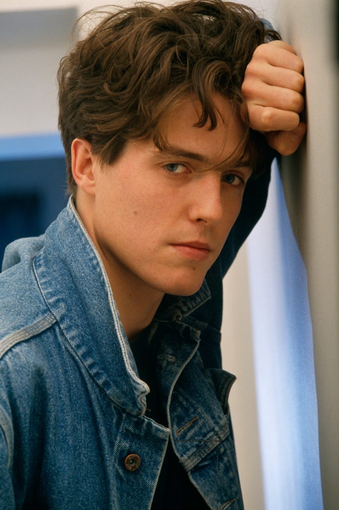 Young Hugh Grant in denim jacket leaning against wall with soft light and intense gaze wallpaper