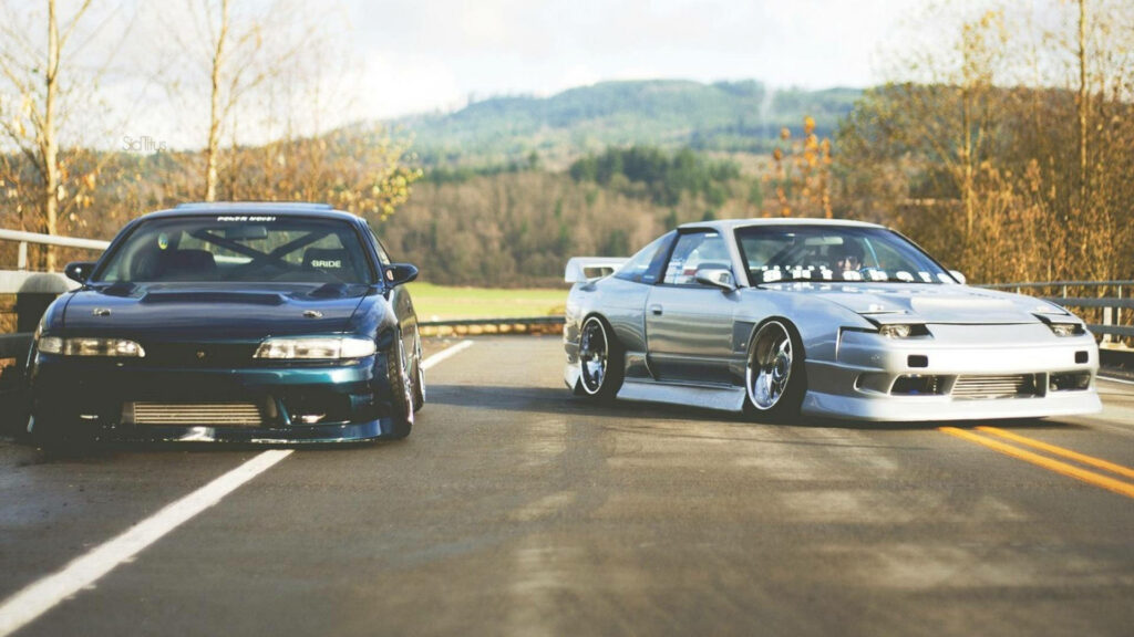 Two classic JDM cars parked on empty road with sleek designs and modified stances in nature Wallpaper