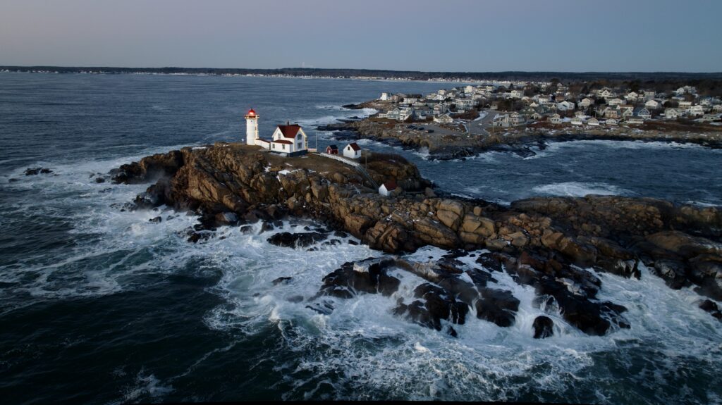 Classic lighthouse on rocky coast with waves and coastal village under soft evening light wallpaper